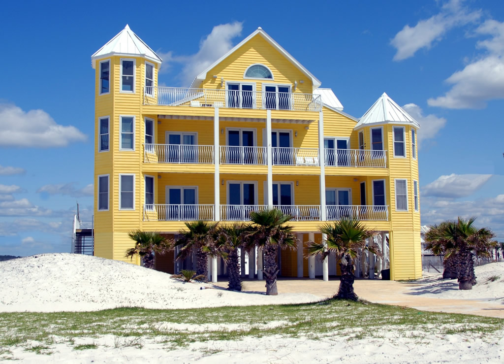 A four story yellow house towers over palm trees on the beach